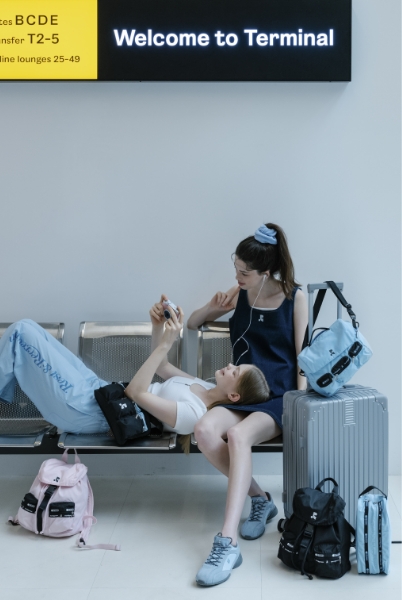 空港の案内板の下のベンチで、寝そべる女性をスマホで撮影する女性