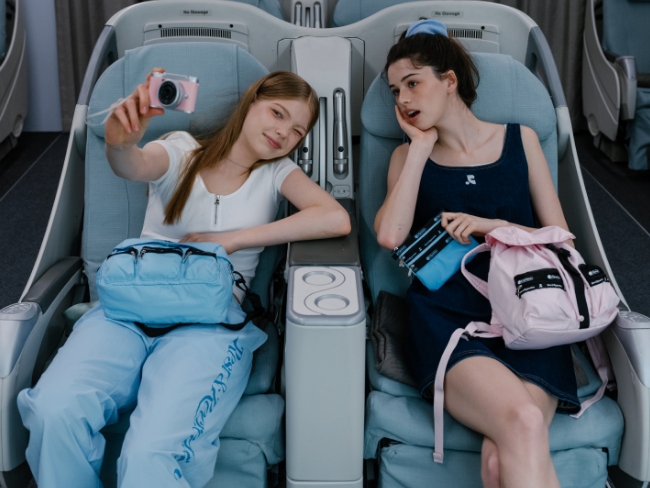 機内の座席で自撮りをする2人の女性と水色とピンクのバッグ